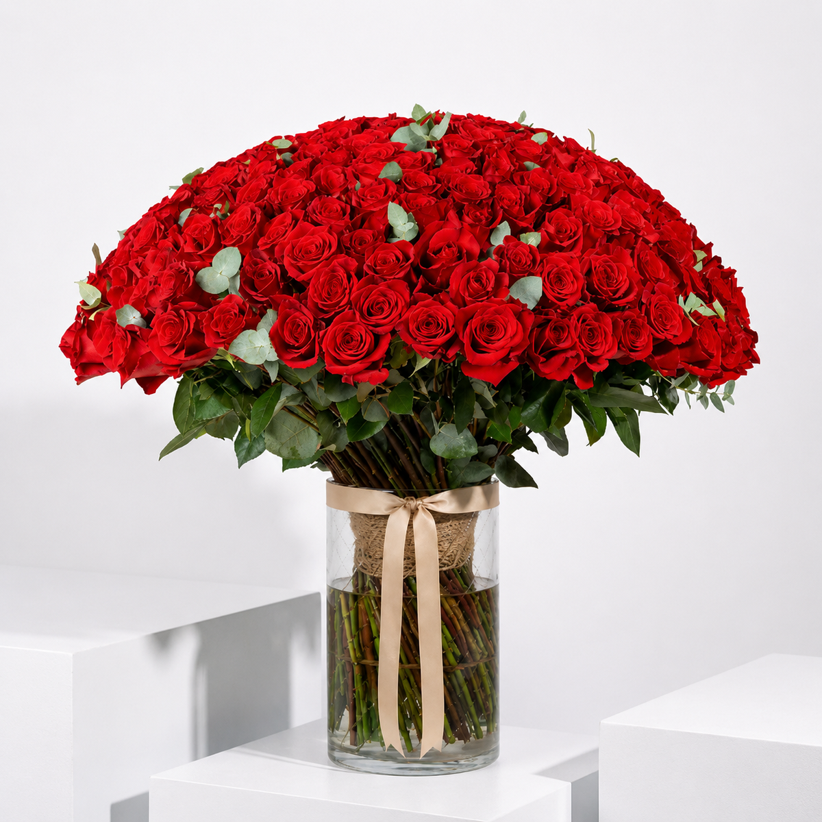 A large bouquet of red roses arranged in a dome shape in a clear glass vase with a beige ribbon